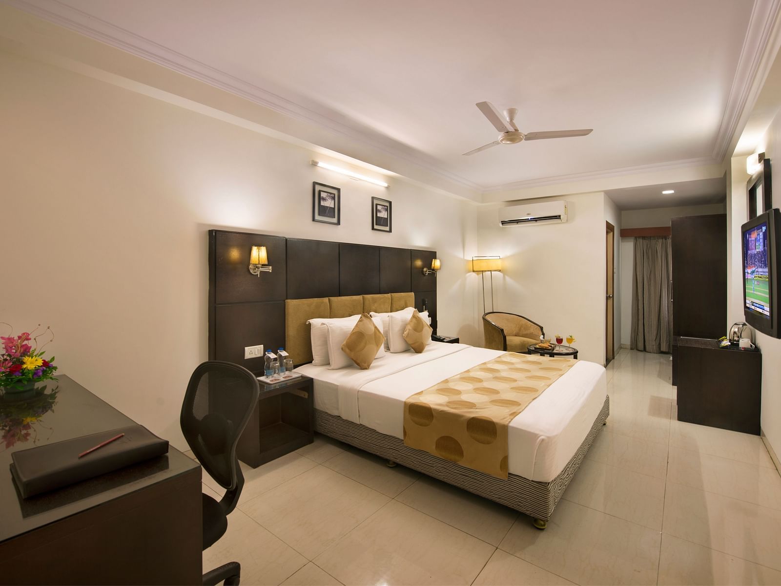 A hotel bedroom with a double bed, dark headboard, a desk and chair, a television, and a seating area with an armchair - Lords Eco Inn Dahej
