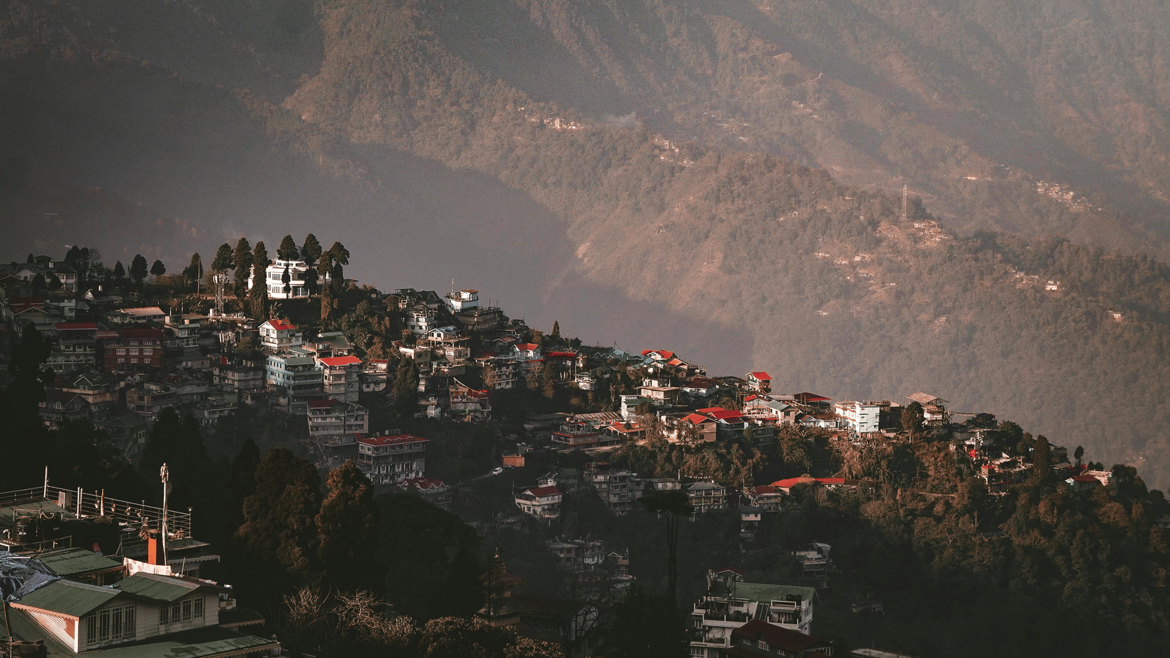 A breathtaking aerial view of a hillside town in Darjeeling, featuring colorful houses nestled among lush green trees against a backdrop of hazy, layered mountains.