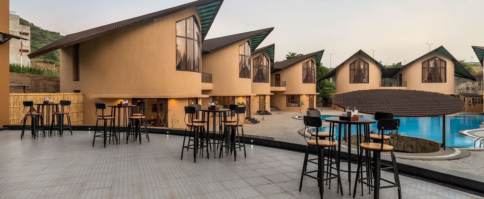 Exterior of Rhythm & Brews at dusk, showcasing modern architecture, outdoor seating areas, and a swimming pool at The Qualia Resort Club & Brewery, Udaipur.