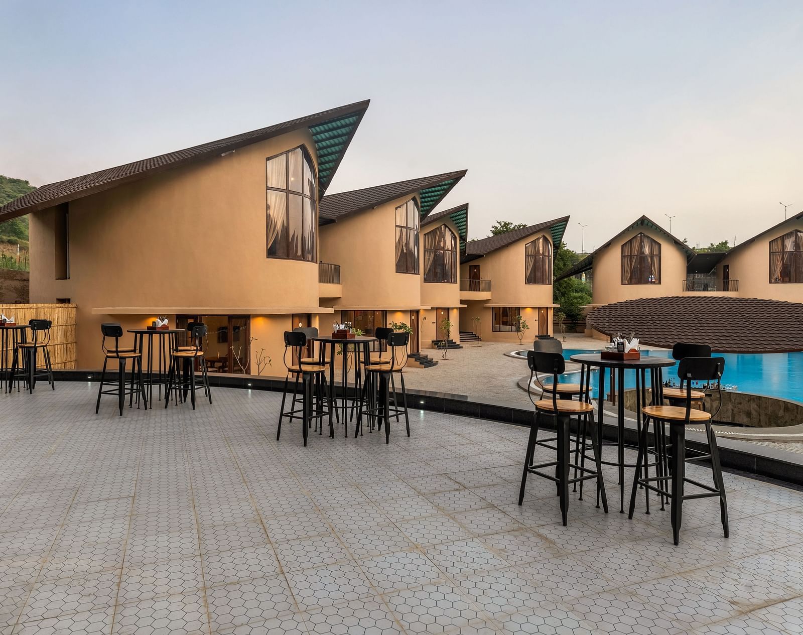 Exterior of Rhythm & Brews at dusk, showcasing modern architecture, outdoor seating areas, and a swimming pool at The Qualia Resort Club & Brewery, Udaipur.