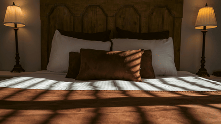 A double bed with multiple pillows and a duvet, positioned between 2 bedside tables with lamps casting light against a wooden headboard.