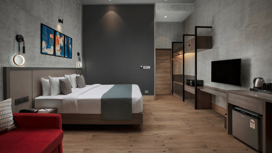 Modern deluxe room at Ocean Palms Goa featuring a king bed with grey tones, wall-mounted TV, wooden flooring, abstract artwork, and a red sofa seating area.