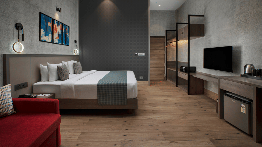 Modern deluxe room at Ocean Palms Goa featuring a king bed with grey tones, wall-mounted TV, wooden flooring, abstract artwork, and a red sofa seating area.