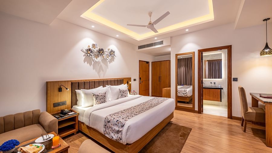 A hotel room with a king size bed, a small couch, and a work desk. It features a unique tray ceiling with lighting, a ceiling fan, and a bathroom with a double vanity visible through an open doorway - Lords Eco Inn Dehradun.