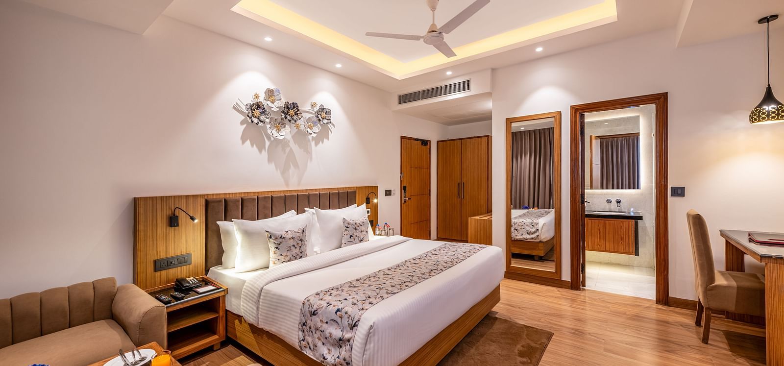 A hotel room with a well-made bed, a beige couch with a side table, a desk with a chair, and an open doorway revealing a bathroom with a double vanity and mirror - Lords Eco Inn Dehradun.