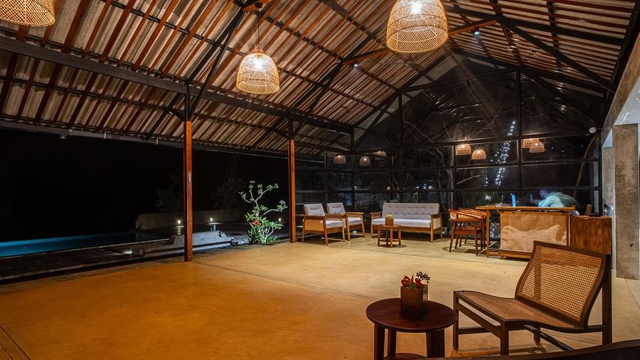 Open-air common area at Vaasta Vagamon with wooden beam ceiling, woven pendant lights, seating, and a pool under the night sky.