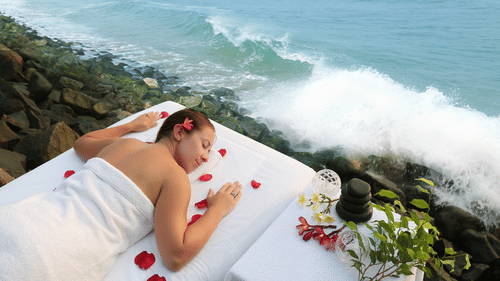 A woman wrapped in a white towel relaxes on a massage table near crashing ocean waves, adorned with red flower petals