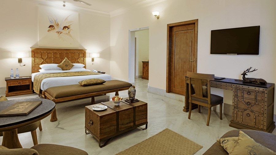 The interior of the spacious plunge pool suite with jacuzzi at  Fateh Safari Suites, Kumbhalgarh featuring a large bed with a carved wooden headboard, a sitting area with a trunk coffee table, a wooden desk and chair, and a wall-mounted television.