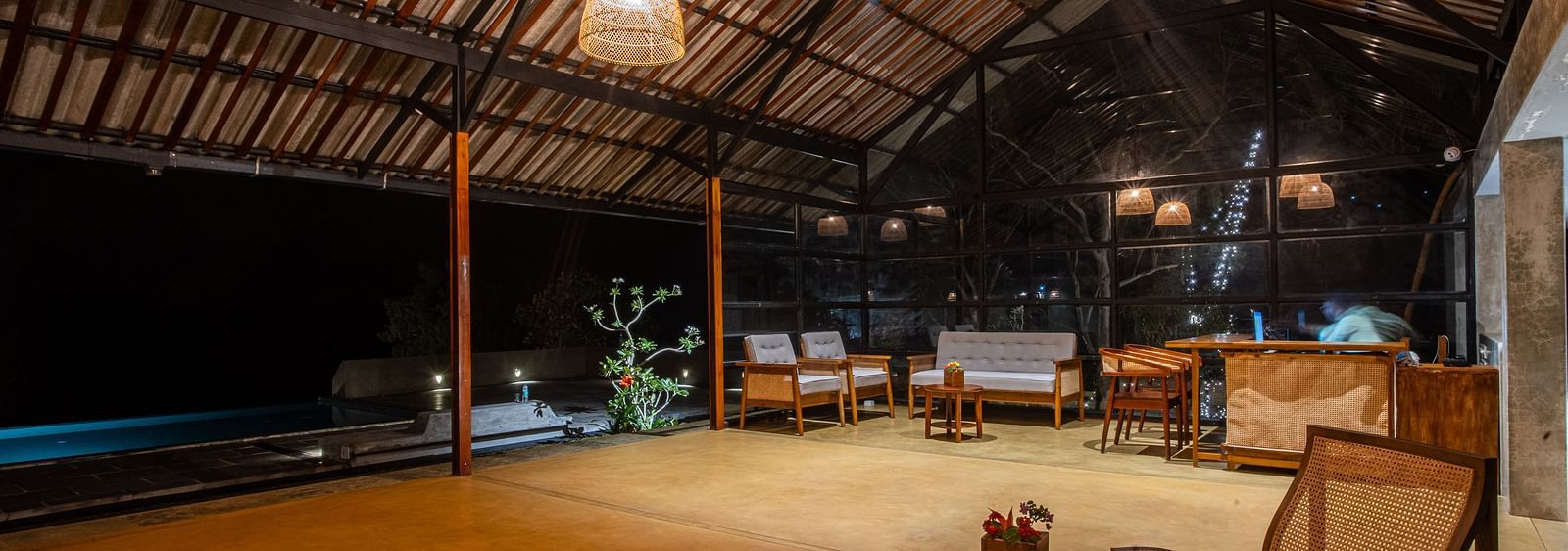 Open-air common area at Vaasta Vagamon with wooden beam ceiling, woven pendant lights, seating, and a pool under the night sky.