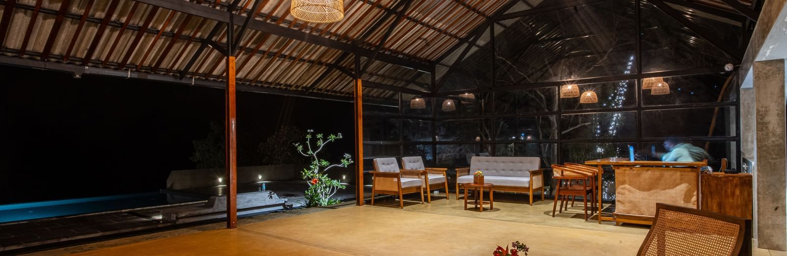 Open-air common area at Vaasta Vagamon with wooden beam ceiling, woven pendant lights, seating, and a pool under the night sky.