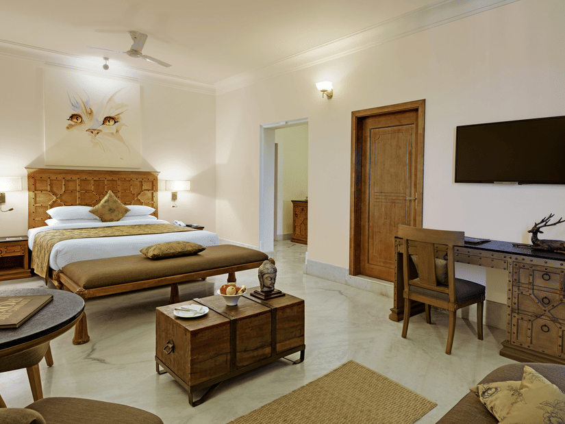 The interior of the spacious plunge pool suite with jacuzzi at  Fateh Safari Suites, Kumbhalgarh featuring a large bed with a carved wooden headboard, a sitting area with a trunk coffee table, a wooden desk and chair, and a wall-mounted television.