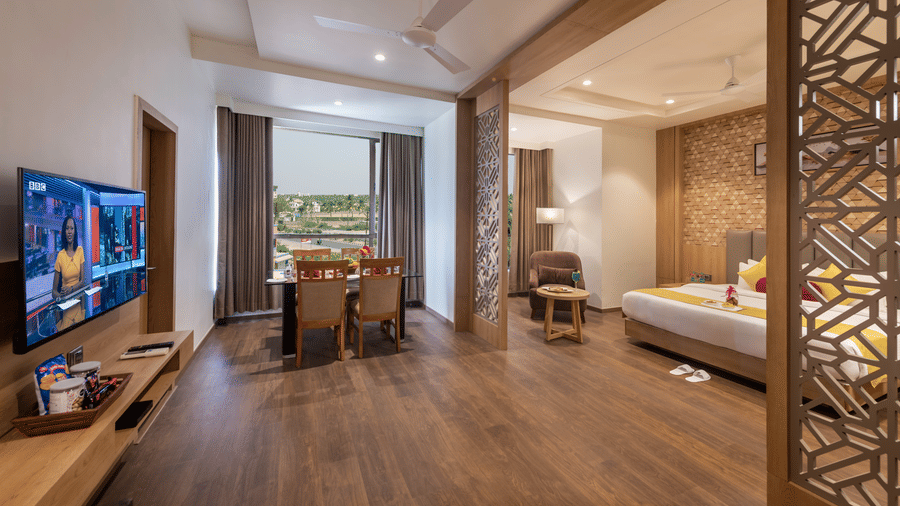 Sitting area of a suite at Lords Inn Somnath, featuring an entertainment unit, and lounge chairs positioned near a large window offering an outdoor view.