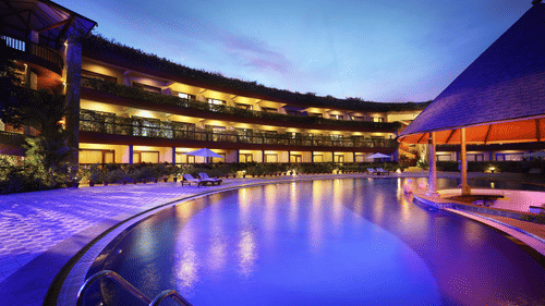 Facade of Uday Suites illuminated against the evening sky, with its glowing pool area and striking curved balconies