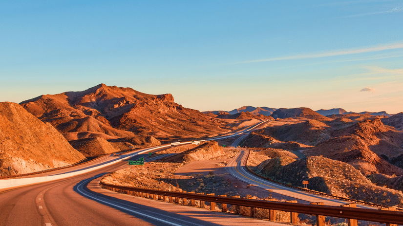 Winding desert highway cutting through rugged, sunlit mountains.