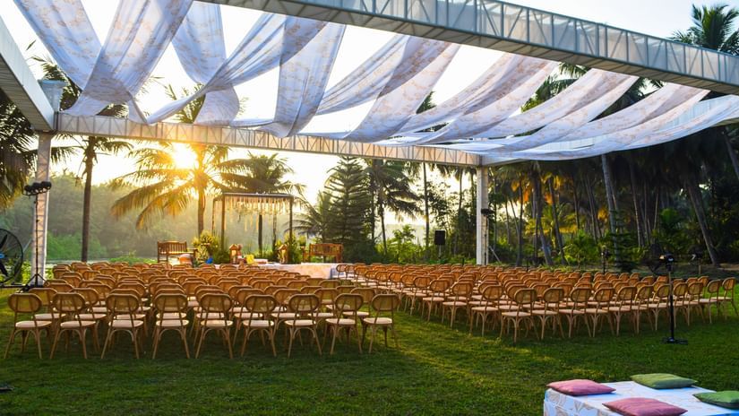 An outdoor event setup featuring rows of empty wooden chairs facing a raised platform under a large canopy, with the sun setting brightly in the background at Ibex River Resort, Pollachi.