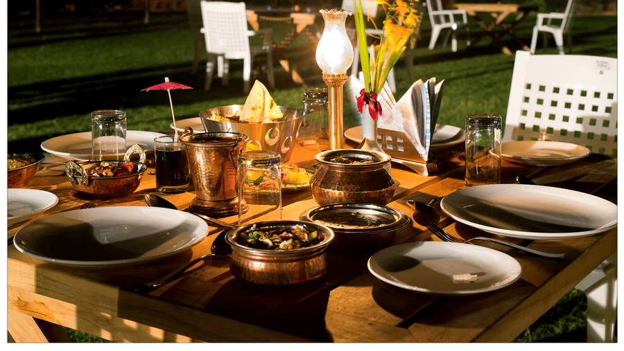 A wooden table outdoors, set with plates, bowls, a lantern, and a floral centrepiece at night at Garh Govind.