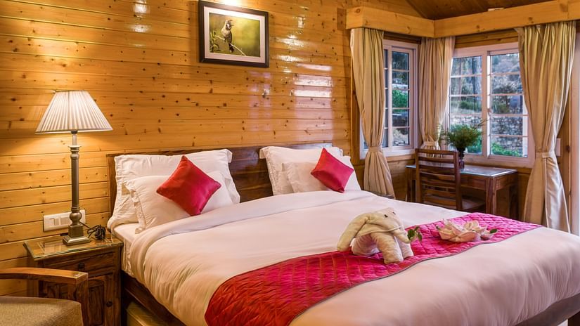  A serene bedroom with light wooden walls and ceiling at Digantaa Resorts in Mukteshwar A large bed with a white comforter and a red decorative runner is prominently featured. A lamp on a bedside table illuminates the left side of the bed