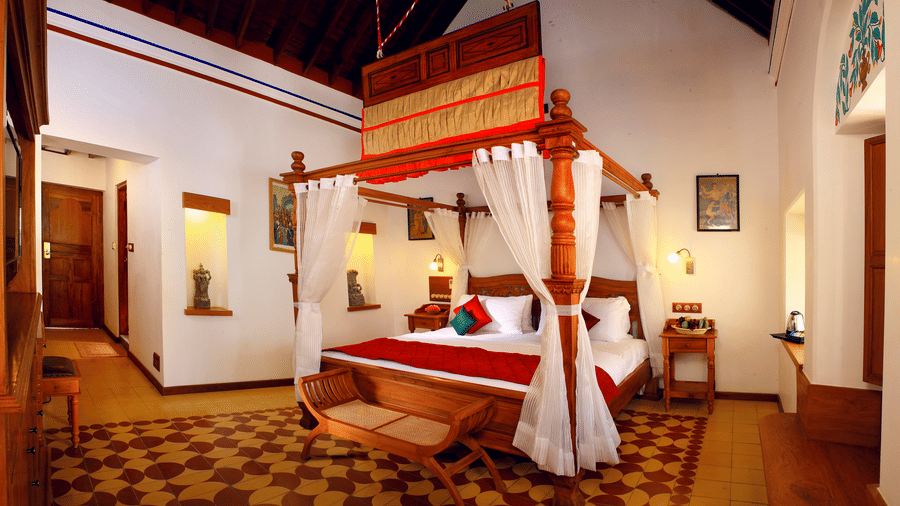 A spacious bedroom at Chidambara Vilas - A Luxury Heritage Resort, with a king size bed, tiled flooring, wooden beams on the ceiling, and natural light from a window.