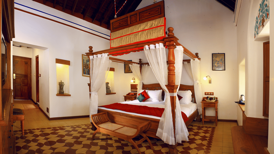 A luxury heritage room at Chidambara Vilas featuring a traditional 4 poster bed with draped canopy, carved wooden accents, classic furnishings, and patterned tile flooring reflecting Chettinad architecture.