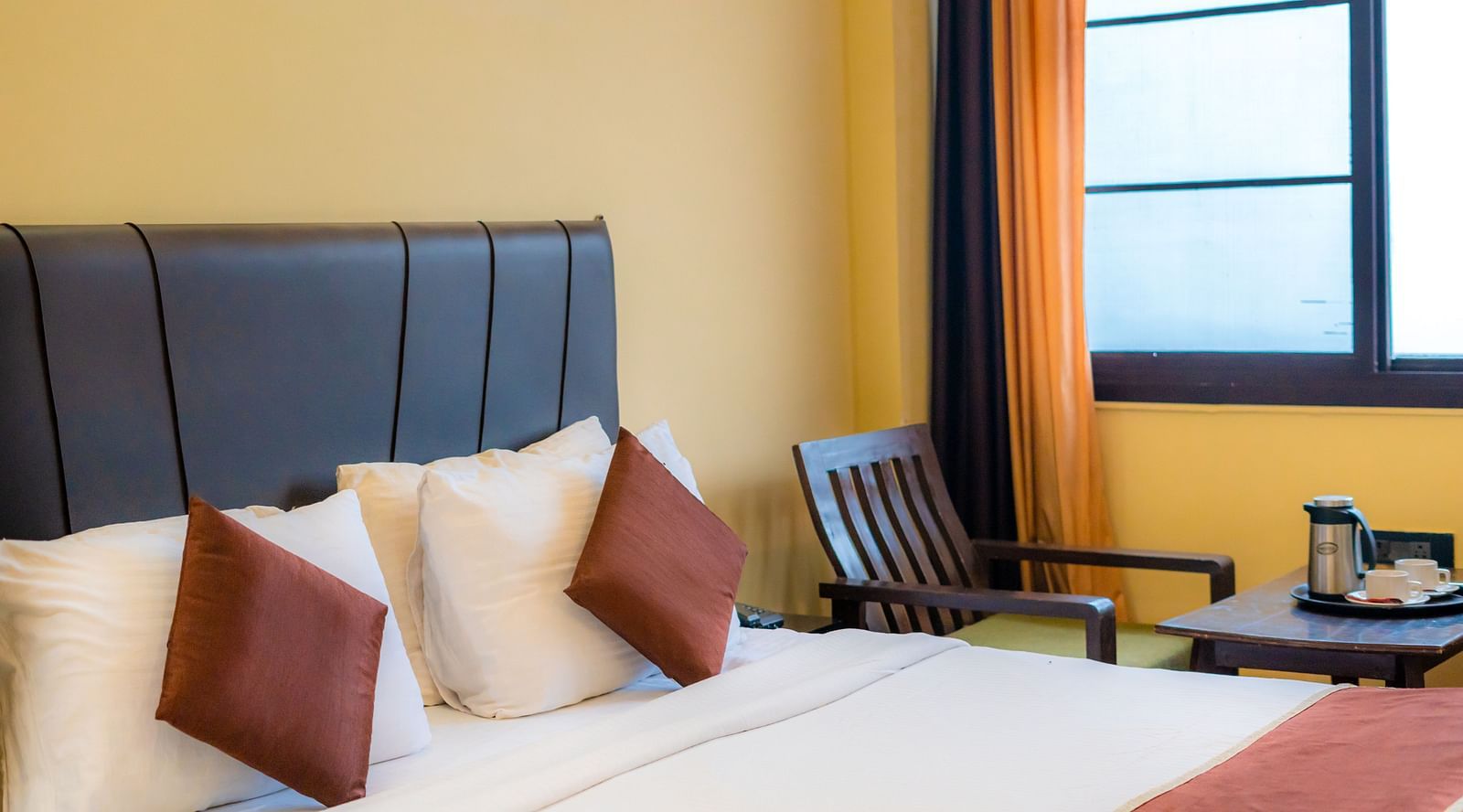 Bright bedroom with a huge bed, a chair and a table, and curtained windows overlooking the valleys at Flutter Hotels and Resorts, Lansdowne.