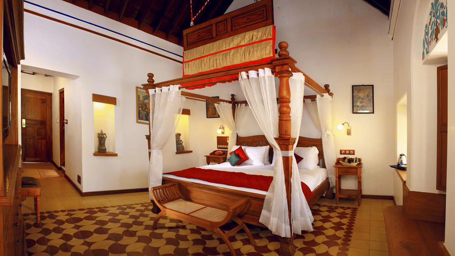 A luxury heritage room at Chidambara Vilas featuring a traditional 4 poster bed with draped canopy, carved wooden accents, classic furnishings, and patterned tile flooring reflecting Chettinad architecture.