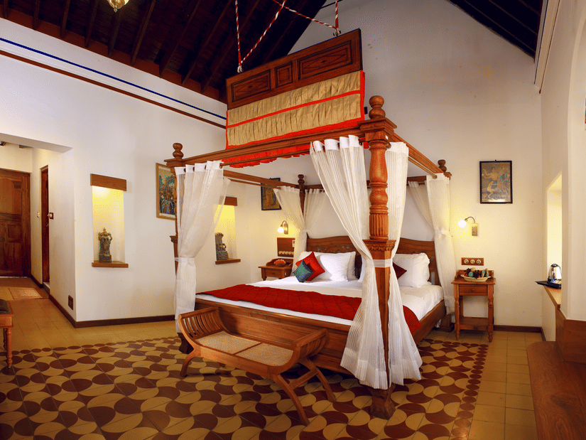 A luxury heritage room at Chidambara Vilas featuring a traditional 4 poster bed with draped canopy, carved wooden accents, classic furnishings, and patterned tile flooring reflecting Chettinad architecture.