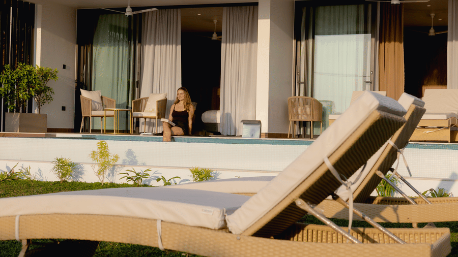 A close-up view of a lounge chair on a lawn at SUKAVI, with a person sitting on the edge of the swimming pool in the background.