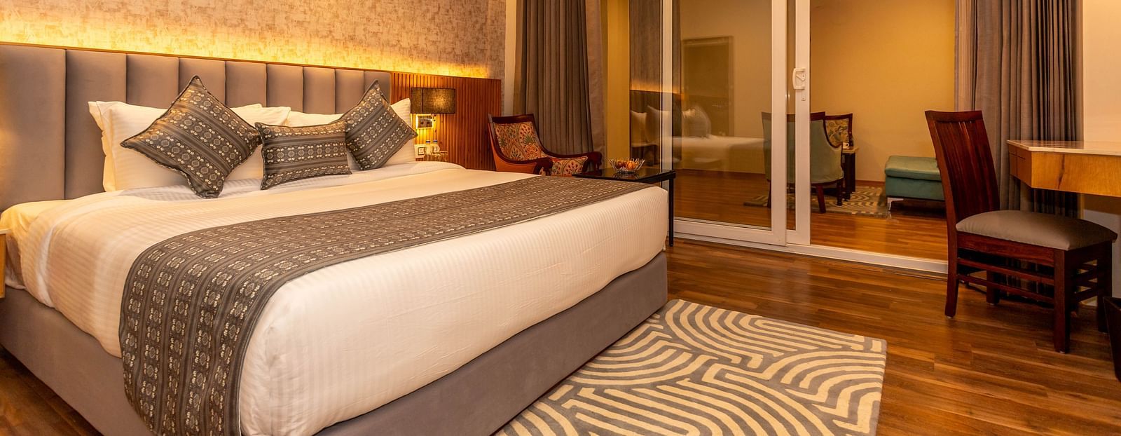 An image of a Family Room with bed, bed side table, lamps, work desk and chair, and an expansive glass door leading to other areas of the room - The Belvedere Himalayan Retreat, McLeodganj