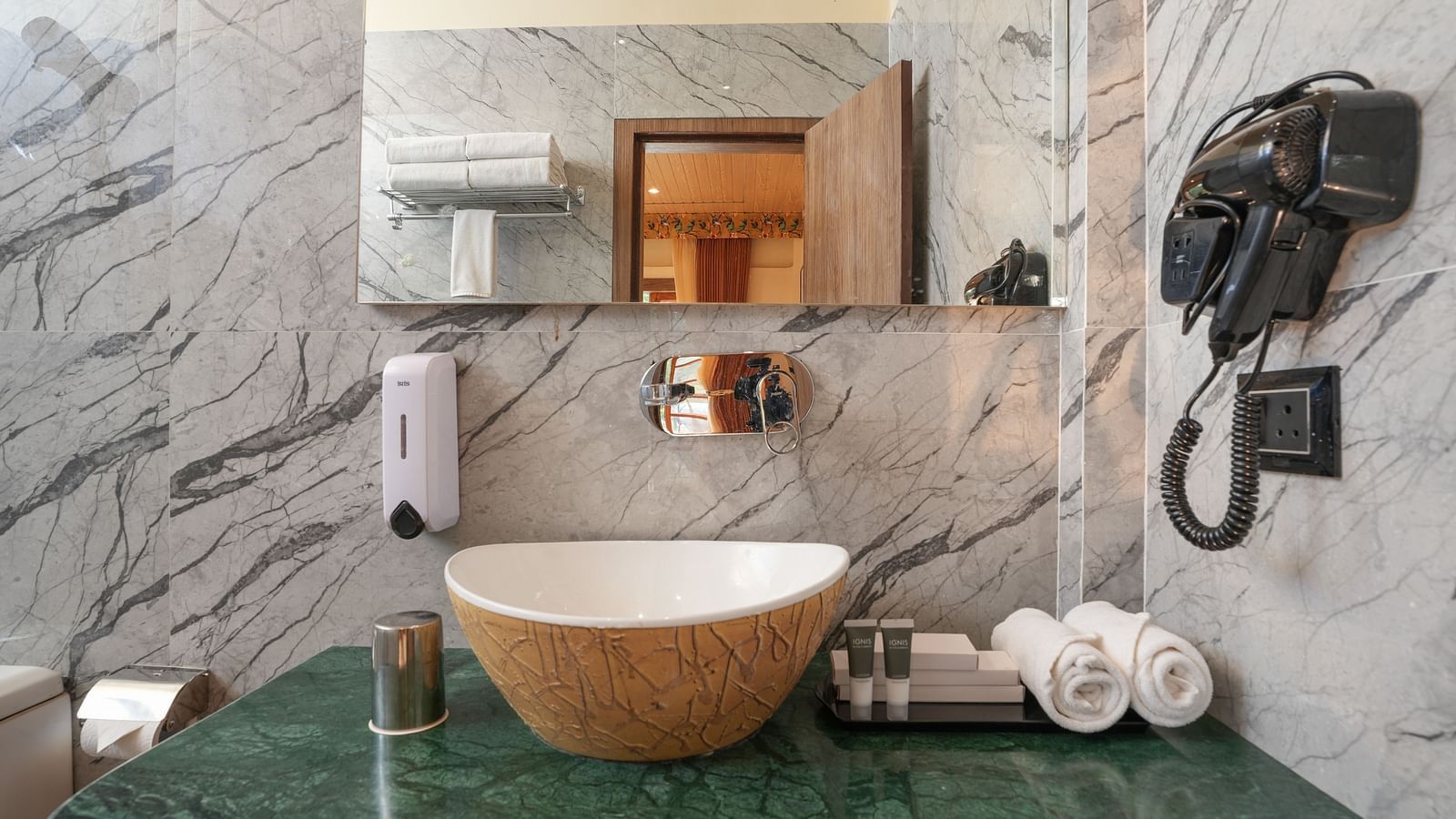 The spacious bathroom of the bungalow features glass separating the shower area, a large washbasin with a mirror, a hairdryer, and wooden storage space underneath at Summit Green Lake Tea Resort & Spa, Kaziranga