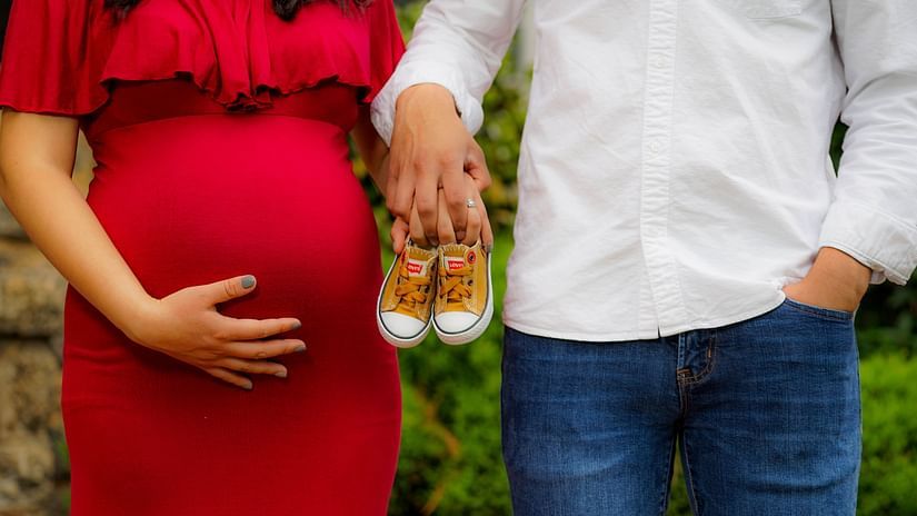 A pregnant woman hold her belly as she stands next to her partner the latter holds up a pair of baby shoes.