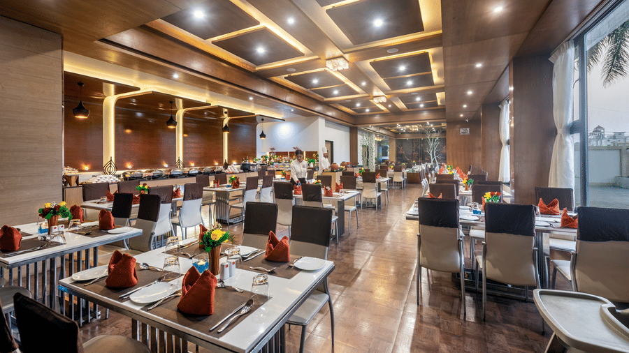A spacious and elegantly designed restaurant with well-set tables adorned with flowers and large windows offering a view of the outdoors at Lords Inn Somnath.