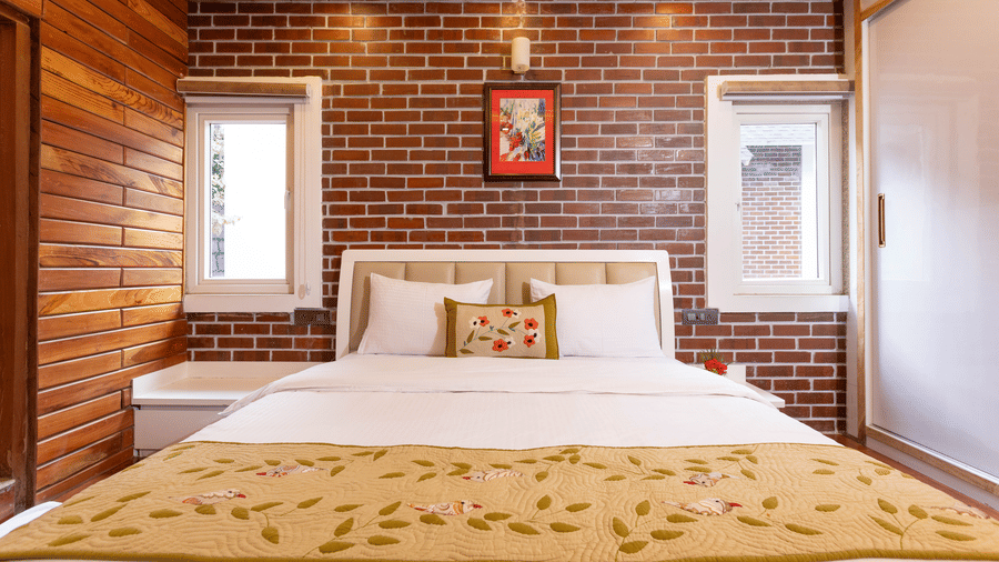 A bright bedroom with a white and gold bedspread, set against a red brick and wooden wall at Megha Resort & Villas, Dehradun.