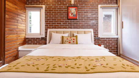 A bright bedroom with a white and gold bedspread, set against a red brick and wooden wall at Megha Resort & Villas, Dehradun.