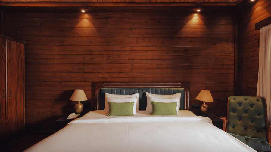 A bedroom at The Ananta Udaipur featuring a king size bed, 2 side tables, bedside lamps, wooden panelled walls, and single armchair.