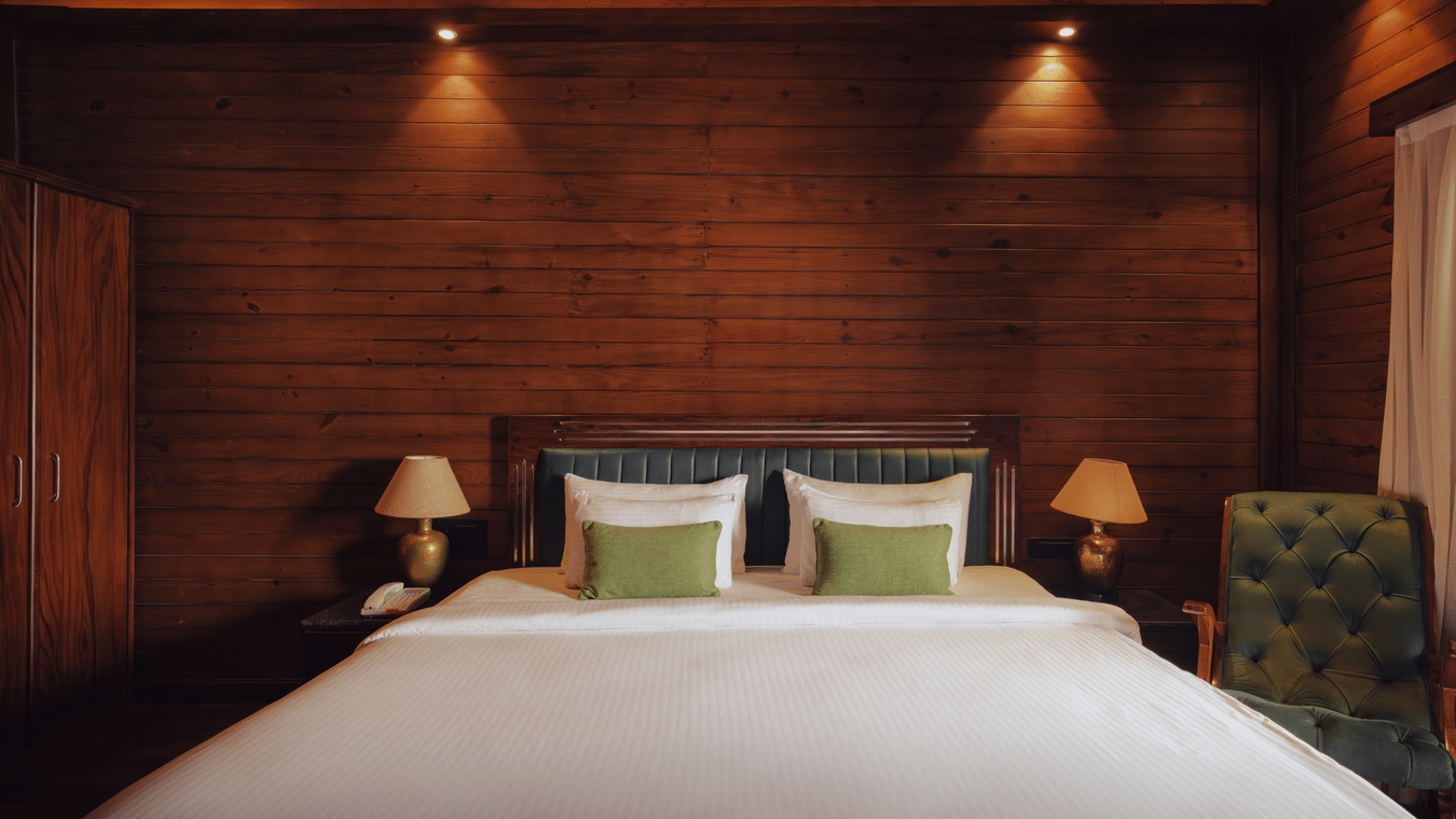 A bedroom at The Ananta Udaipur featuring a king size bed, 2 side tables, bedside lamps, wooden panelled walls, and single armchair.