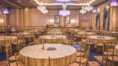 A spacious, elegantly decorated ballroom is set with numerous round tables and gold chairs, ready for an event.