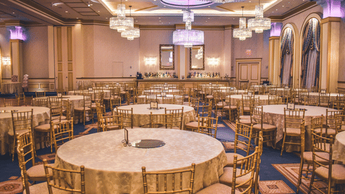 A spacious, elegantly decorated ballroom is set with numerous round tables and gold chairs, ready for an event.