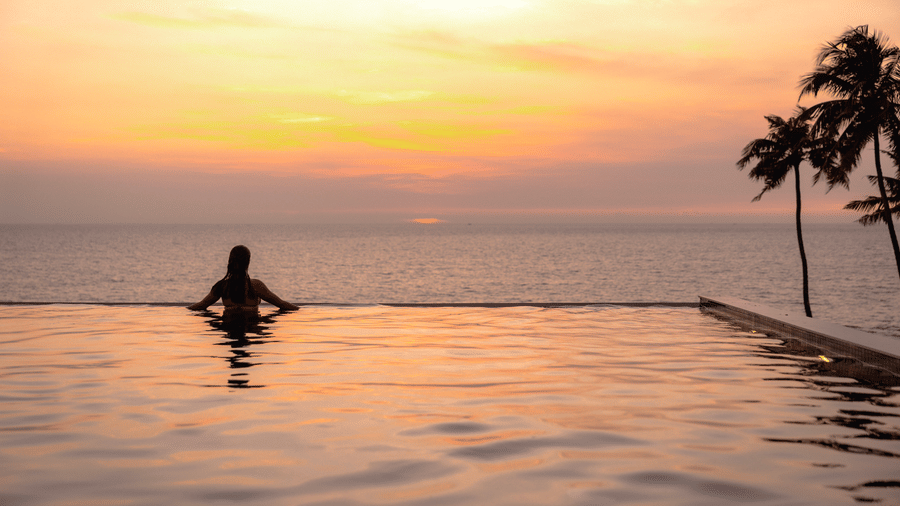 A person partially submerged in the infinity pool at SUKAVI, gazing towards the ocean beneath a sunset sky, with palm trees framing the scenic coastal view.