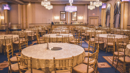 A grand ballroom features large round tables and gold chairs beneath ornate, glittering chandeliers