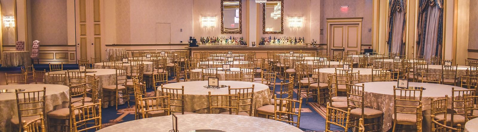 A luxurious banquet hall with round tables, gold chairs, elegant chandeliers, and soft ambient lighting, set up for a formal event.