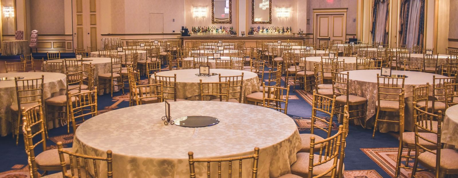 A luxurious banquet hall with round tables, gold chairs, elegant chandeliers, and soft ambient lighting, set up for a formal event.