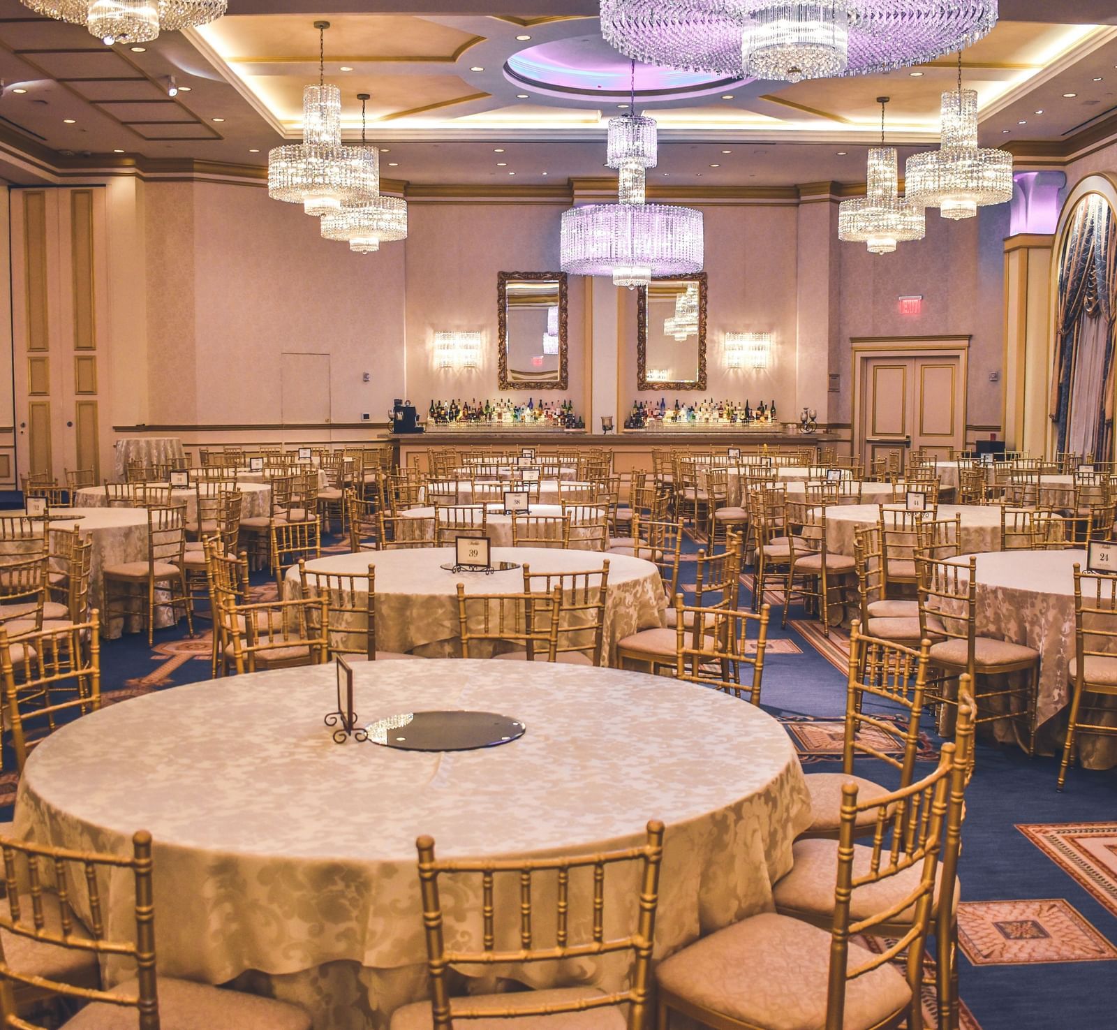A brightly-lit banquet hall with chairs surrounding various round tables in the hall featuring beautiful chandeliers hanging from the ceiling.