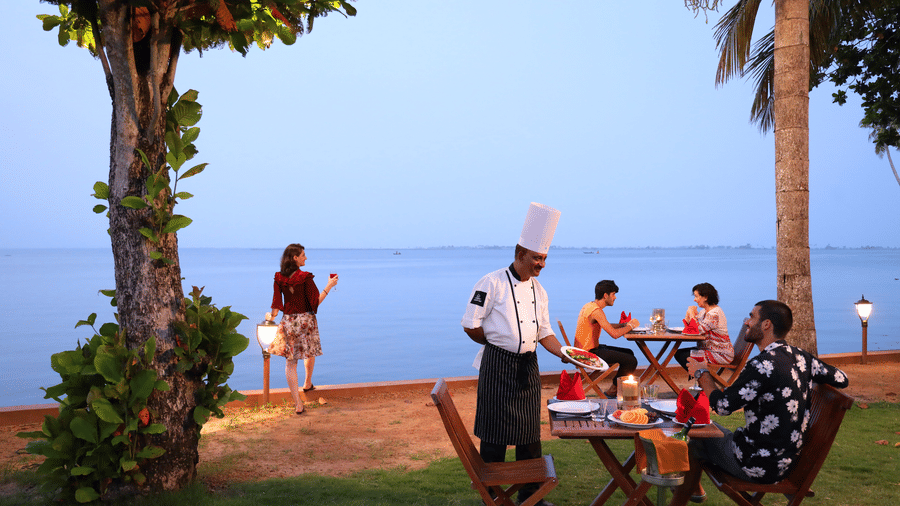 Outdoor seaside dining with a chef serving guests, a woman admiring the ocean, and a tranquil evening atmosphere under palm trees at Uday Backwater Resort