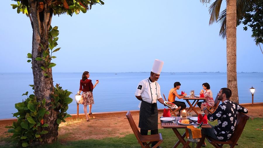 Outdoor seaside dining with a chef serving guests, a woman admiring the ocean, and a tranquil evening atmosphere under palm trees at Uday Backwater Resort