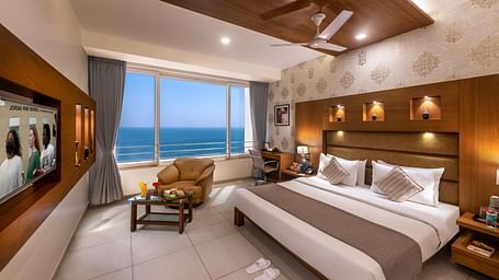 A room with a bed, seating area facing a large window with an ocean view, and a workspace.