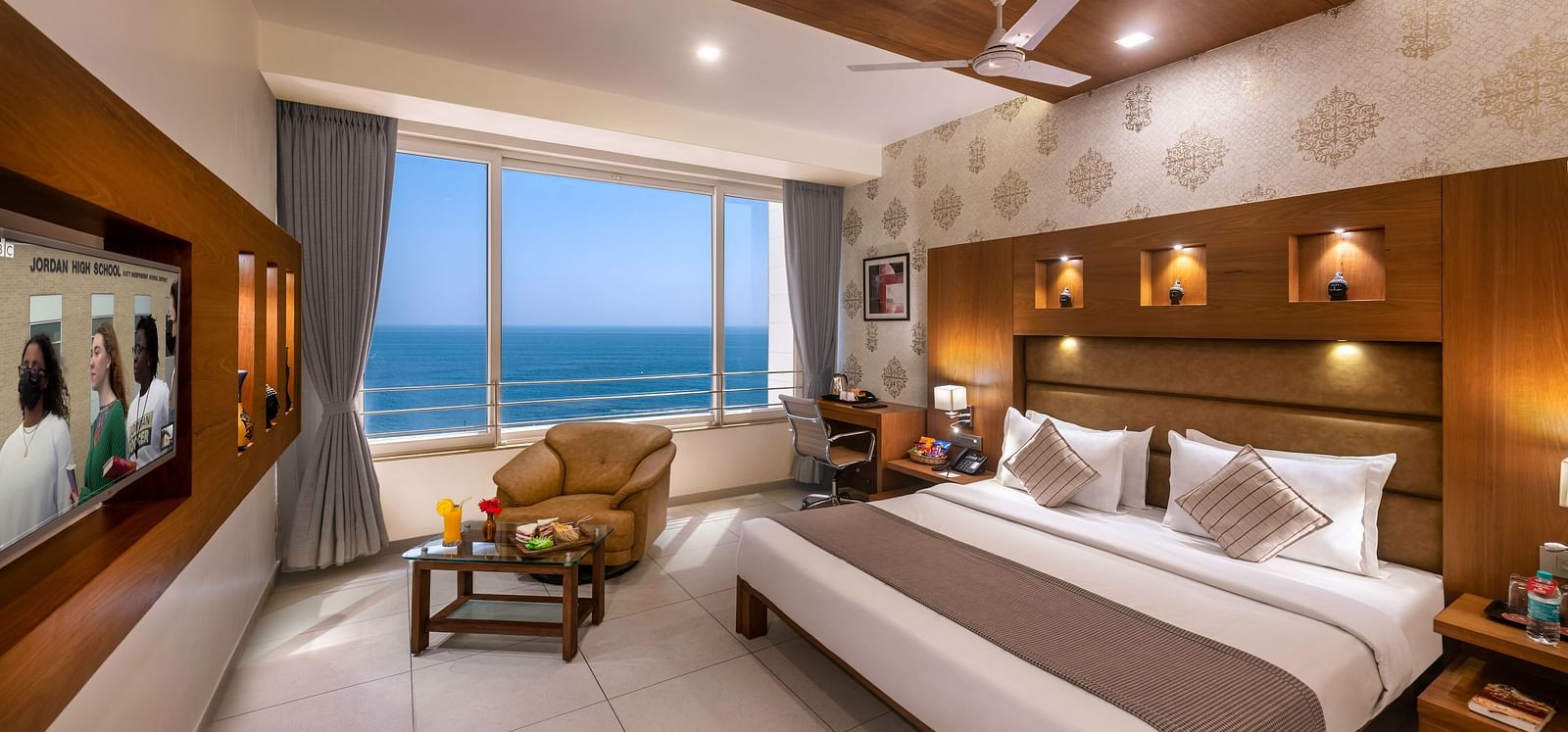 A room with a bed, seating area facing a large window with an ocean view, and a workspace.