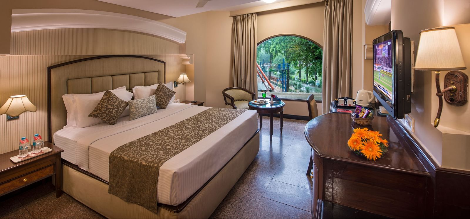 Hotel room with a neatly made bed, a decorative lamp, and a view of greenery through a window - Lords Inn Vadodara