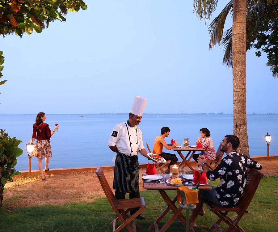 Outdoor seaside dining with a chef serving guests, a woman admiring the ocean, and a tranquil evening atmosphere under palm trees at Uday Backwater Resort