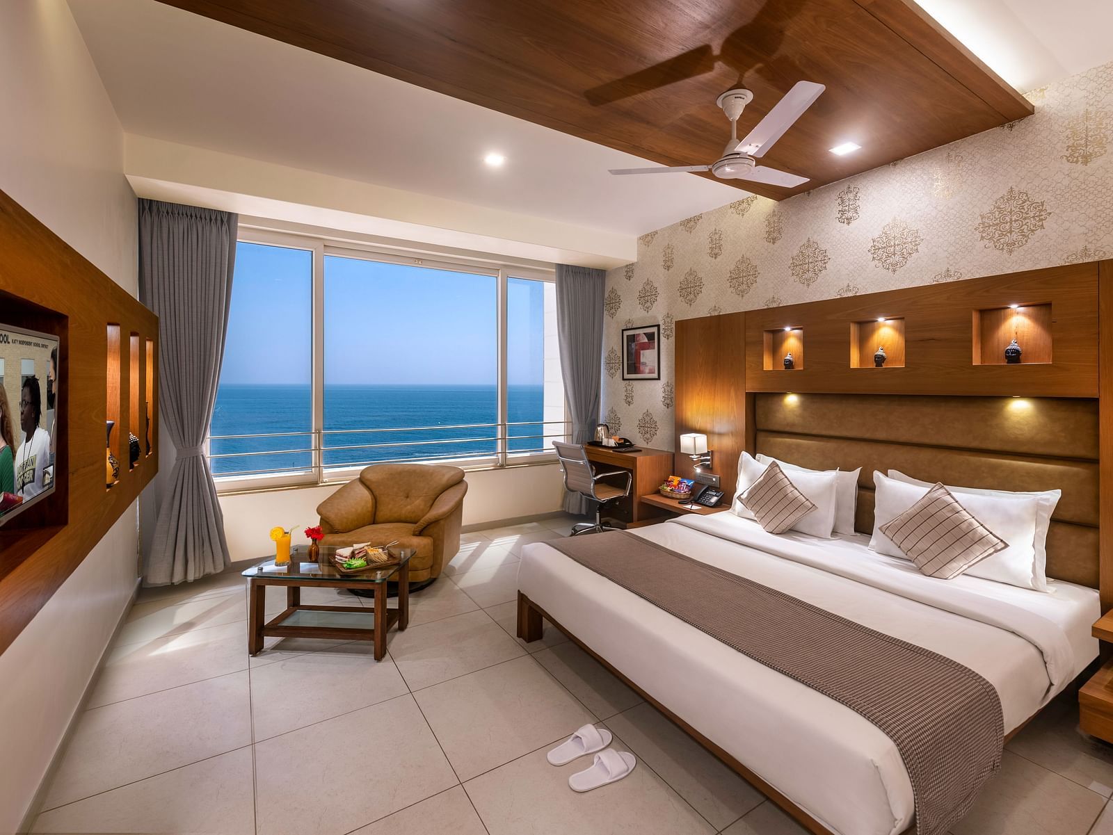 A room with a bed, seating area facing a large window with an ocean view, and a workspace.