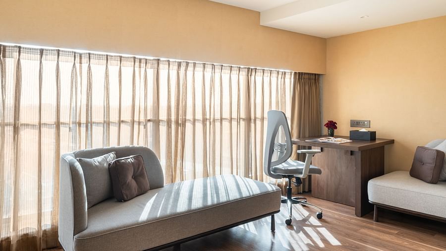 Bright corner of a room with floor-to-ceiling curtains, modern chaise lounge, and a work desk with an ergonomic chair at Clarks Inn Suites, Hyderabad